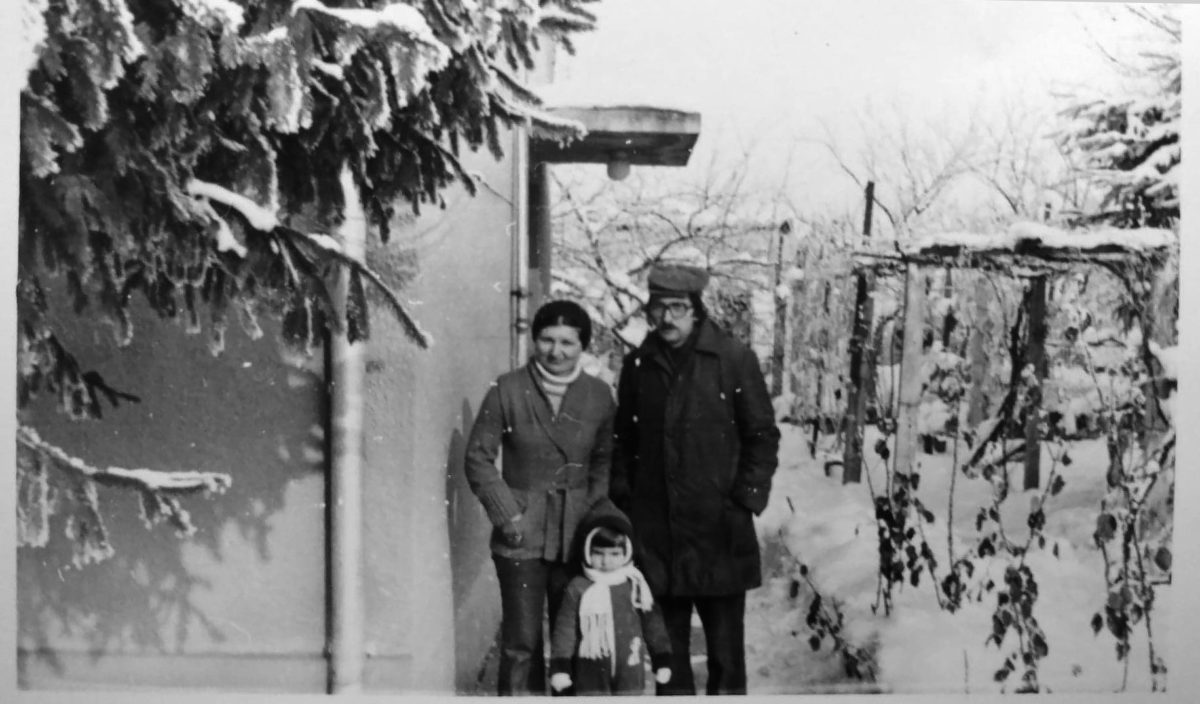 black and white image of three people in front of a building. lots of snow on the ground and on the tree branches.